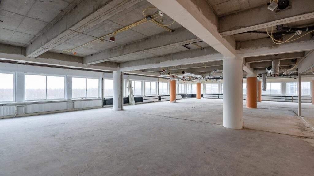 Leegstaand en onafgewerkt kantoorpand aan het Koningin Wilhelminaplein met betonnen vloeren, zichtbare leidingen en grote ramen.